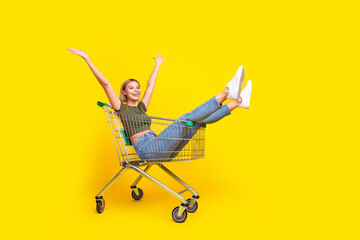 Full size photo of good mood girl dressed khaki crop top sit in shopping cart raising hands up isolated on yellow color background
