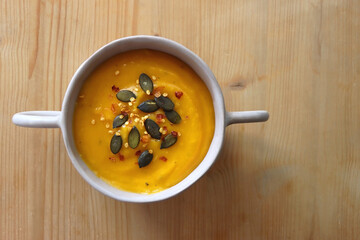 Bowl of creamy pumpkin and potato soup, with pumpkin seeds and chilli flakes toppings. Top view, wooden background.