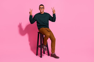 Full length photo of handsome good mood guy wear sweatshirt sitting bar chair showing two v-signs emtpy space isolated pink color background