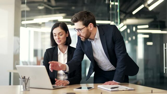 Boss consult young female worker at workplace in business office. Male manager in suit advises, talking to an employee about a development project. Executive explains work issues using laptop computer