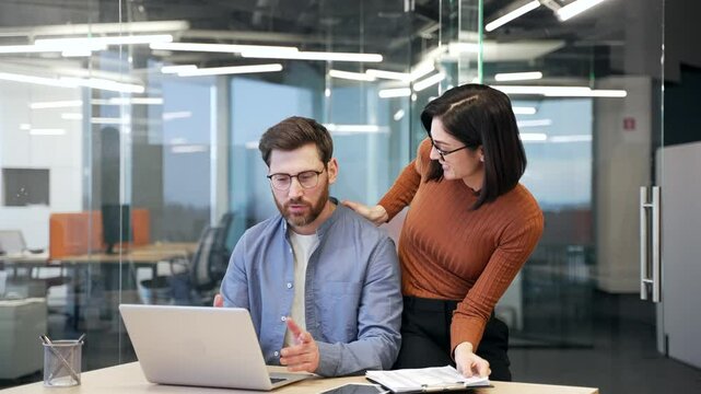 Young woman sexually harasses a male coworker places hand on shoulder at workplace in business office. Beautiful female worker flirts with a shocked and concerned male colleague who is sitting at desk