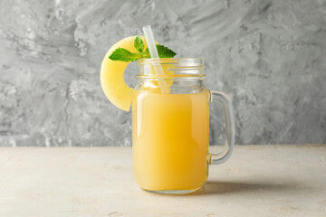 Tasty pineapple juice in mason jar, mint and slice of fresh fruit on grey textured table, closeup