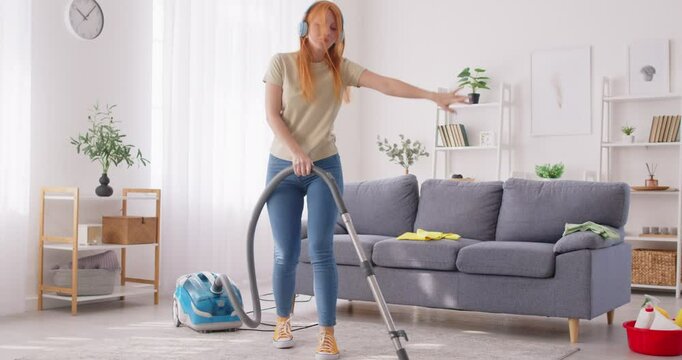 Carefree funny happy young woman vacuuming, teen girl cleaning house, living room having fun dancing with, music in ear headphones overjoyed millennial feels excited to enjoy making chores, entertain 