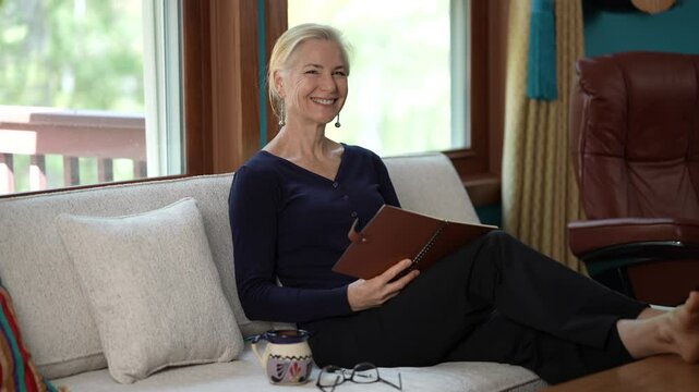 Portrait pf happy mature blonde woman with journal on sofa in living room writing and thinking of idea.