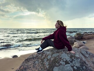 Woman in Burgundy Hoodie Sitting on Rock by the Sea on a Cloudy Day