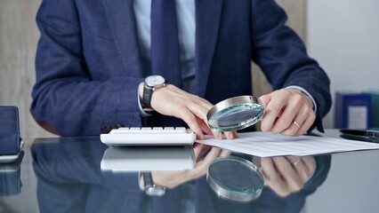 Auditor man analyzing financial documents with magnifying glass and calculator at his glass office desk in formal blue jacket. Business people concept
