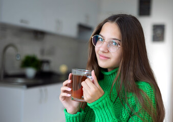 Young girl with glasses enjoying tea in the kitchen. Young girl with long brown hair wearing glasses and a green knitted sweater holding a clear glass cup of tea in a modern kitchen.