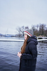 beautiful young girl with long blond hair in a hat and a long down jacket on the pier of a winter lake