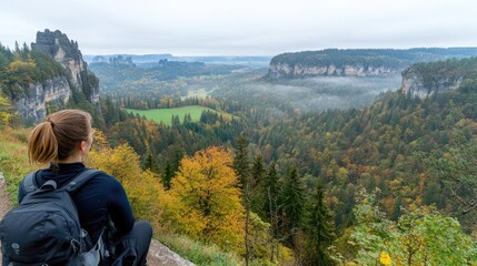 A woman stands in an autumn forest, admiring a beautiful mountain view with a waterfall. She wears a brown hat and touches her backpack, embracing the peaceful atmosphere