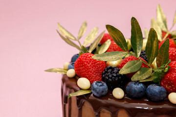A beautifully decorated chocolate cake features an assortment of vibrant strawberries, blueberries, blackberries, and golden embellishments, set against a soft pink backdrop