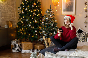 Cozy holiday moment of a young woman in a red sweater and Santa hat communication enjoying her phone amidst festive decorations and Christmas trees