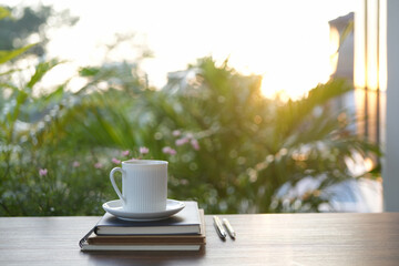 White coffee cup and notebook with lush green plant on wooden table under sunlight in the garden