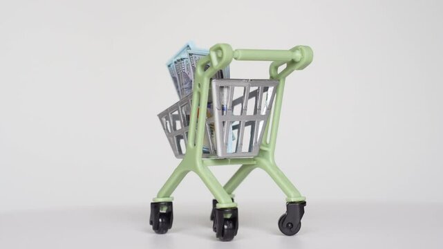 Mini shopping cart overflowing with toy currency on a white background, representing concepts of finance, shopping, and consumerism