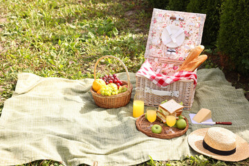 Picnic basket with tableware, different snacks, juice and hat outdoors, space for text