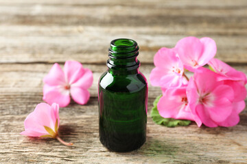 Bottle of geranium essential oil and beautiful flowers on wooden table, closeup