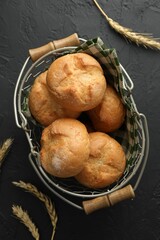 Metal basket with homemade tasty buns and spikelets on black textured table, flat lay