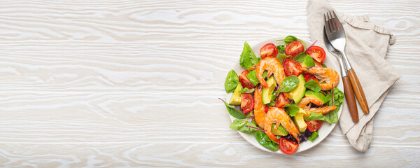 Healthy salad with grilled shrimps, avocado, cherry tomatoes and green leaves on white plate with cutlery on white wooden rustic background top view. Clean eating, nutrition and dieting, copy space..