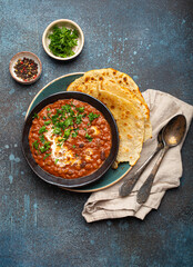 Traditional Indian Punjabi dish Dal makhani with lentils and beans in black bowl served with naan flat bread, fresh cilantro and two spoons on blue concrete rustic table top view.