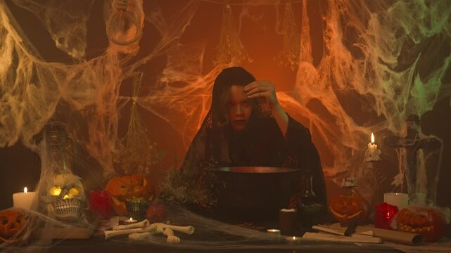 A witch in black clothes cooking a potion in a scary kitchen with pumpkins and a human skull and bones.