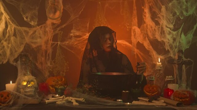A witch in black clothing brews a potion in a scary kitchen with pumpkins and human bones and bones.