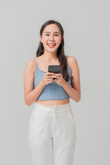 Happy young woman holding smartphone, wearing light blue crop top and white pants, exuding confidence and joy. She stands against neutral background, ready to connect