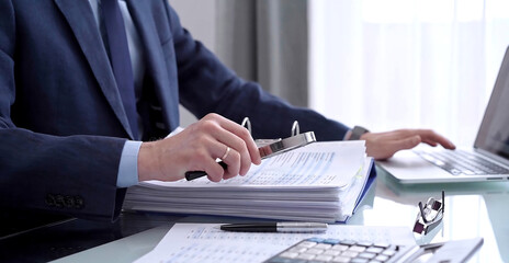 Businessman using a laptop computer and magnifying glass while analyzing financial documents at desk in modern office. Audit and taxes in business