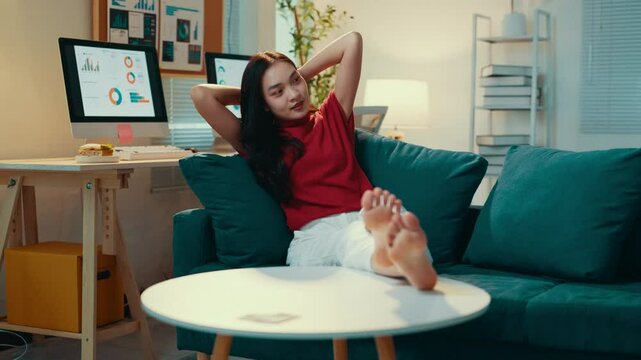 Young woman enjoys a moment of relaxation on her couch after a long day of work. With her feet up on the coffee table, she stretches and smiles