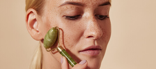 Close-up portrait of a young woman with freckles using a jade roller. Blond female doing beauty...