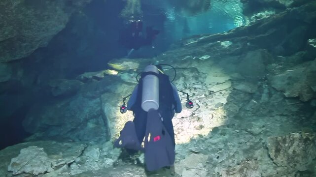 A man in a black wet suit is diving into a cave. The cave is dark and the water is murky