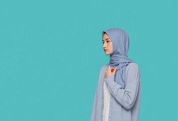 Fototapeta premium A woman in a soft blue hijab and matching cardigan gazes thoughtfully to the side, standing against a turquoise backdrop. Her calm demeanor reflects simplicity and elegance. Generative AI