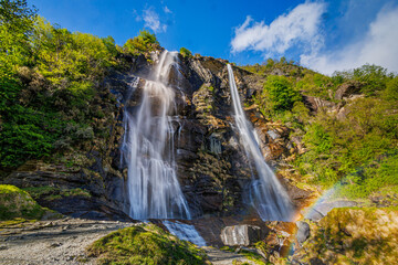 A majestic double waterfall plunges down rugged cliffs, with a rainbow shimmering at the bottom. Bright foliage and clear skies enhance the scenic beauty of this natural wonder
