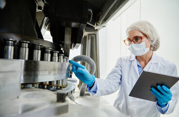 Professional female process technician in vinyl gloves and mask inspecting machinery while pointing...
