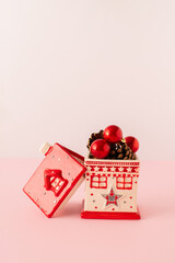 Small ceramic house full of Christmas decoration balls and pine cones with red roof beside, pink background Holiday background concept. 
