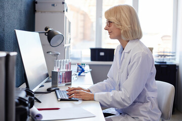Side view of positive female lab chemist typing on computer keyboard while studying chemicals...