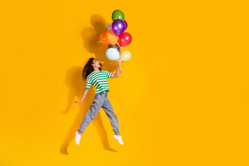 Full size photo of pretty young girl jump fly balloons wear striped t-shirt isolated on yellow color background