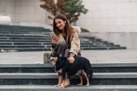 Businesswoman petting dog and using smartphone on city street