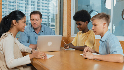 Fototapeta premium Smart girl ask teacher a question while show idea while diverse student working classwork. Group of multicultural children pointing at notebook while happy boy sharing idea at classroom. Edification.