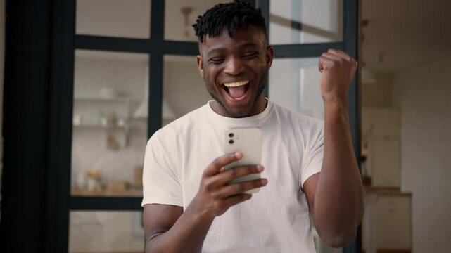 African American ethnic happy excited guy cheerful man husband homeowner male winner user read mobile phone message luck win online betting gambling addict cellphone victory rejoice celebrate at home