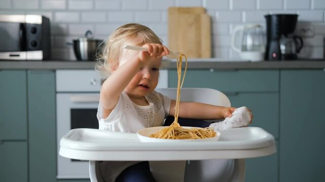 Charming baby sits in a high chair, concentrating on eating pasta bolognese with a fork. The cozy kitchen provides a warm backdrop to this delightful lunchtime