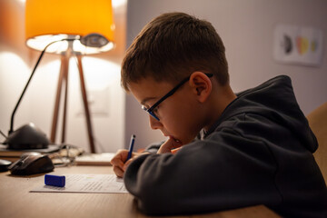 a young boy wearing glasses, deeply focused on writing or doing homework at a desk under warm lighting, with a lamp and study materials scattered around, creating a cozy and concentrated atmosphere.