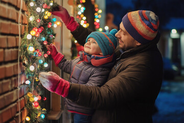 Man with his small son hangs New Year's garlands with lights on the entrance of the house from the outside, on the street.