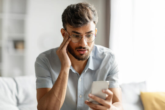Shocked indian guy looking at cellphone screen, sitting on couch at home, holding phone and touching his head, reading bad news or message, copy space
