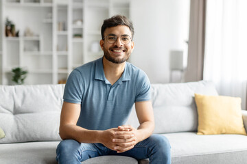Obraz premium Portrait of handsome indian man resting at home alone, sitting on couch in cozy living room and smiling at camera, cheerful hindu guy in casual outfit, free space