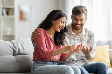 Happy indian spouses holding positive pregnancy test, sitting on couch at home together, couple...