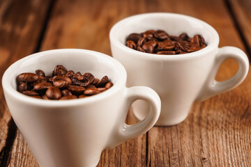 Two white ceramic cups with roasted coffee beans inside on brown wooden table. Coffee shop, caffeine, roast concept. Roasted beans for mass market sale, horizontal photo.