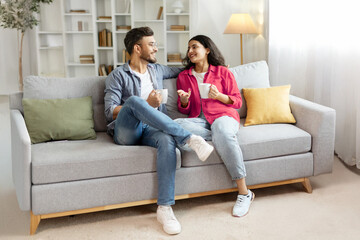 Happy indian couple relaxing and drinking coffee, sitting on couch at home, talking , free space