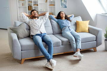 Obraz premium Joyful hindu couple lounges on sofa, hands behind their heads, enjoying leisurely moment in living room interior at home setting