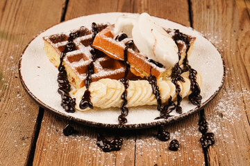 Sweet dessert waffles. Belgian waffles with banana, ice cream and chocolate on top. Delicious dessert for tea time. Served with ice cream. Plate of waffles with ice cream and banana on wooden table.