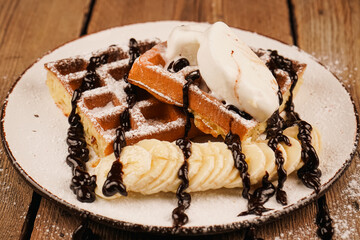 Delicious dessert for tea time. Belgian waffles with banana, ice cream and chocolate on top. Sweet dessert waffles. Served with ice cream. Plate of waffles with ice cream and banana on wooden table.