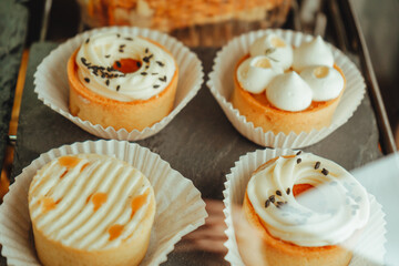 Display case with a variety of cakes, cupcakes and deserts. The tarts with mango and tarts with dork cheese are arranged in a row on black plate.